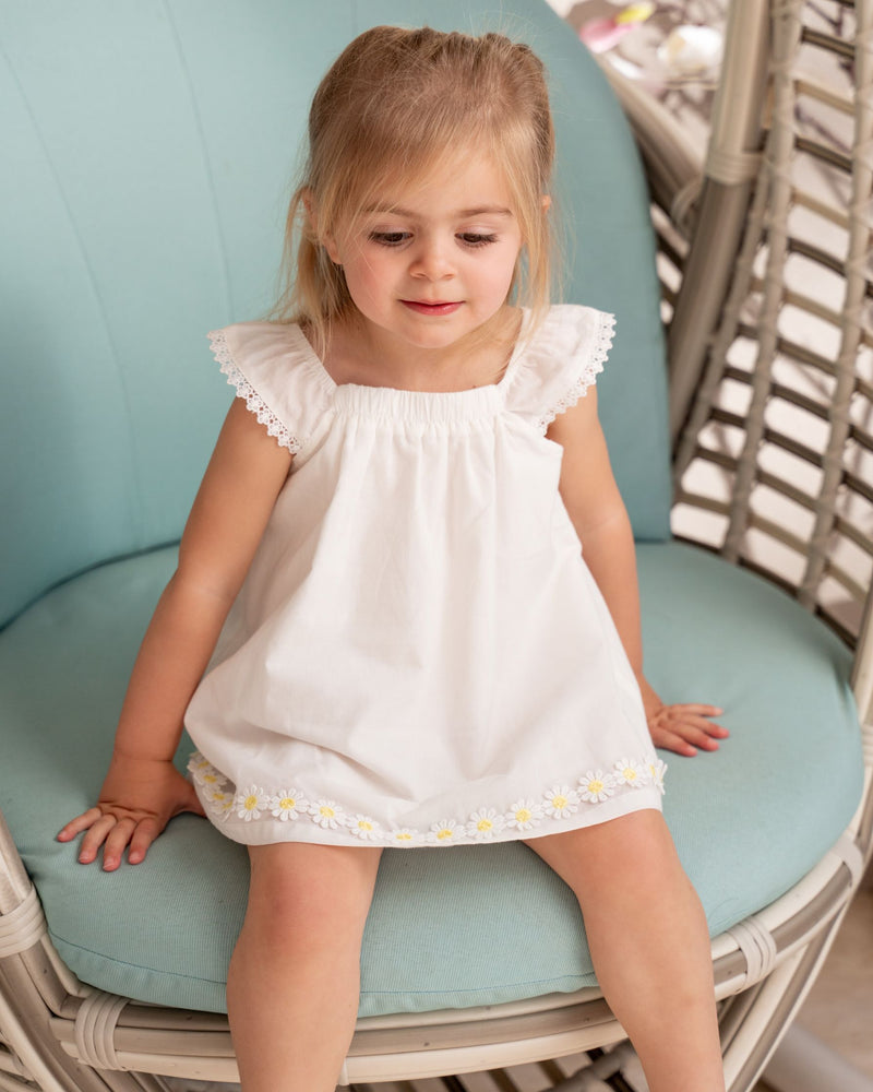 Little girl sitting on the swing chair wearing Organicline sleeveless natural white dress, featuring angle-sleeves with a line of lace eyelet on the edge and a gentle elastic neckline. Small yellow sunflowers embroidered at the end of the hem making this clean and simple dress a playful style. 